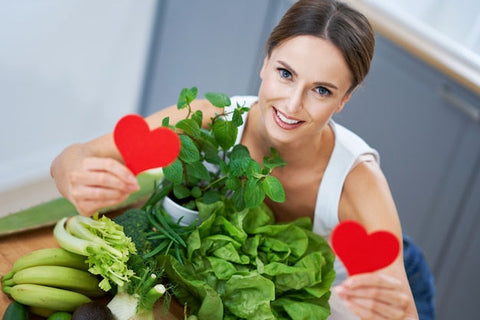 Frau mit Salat, Bananen und Kräutern in Küche hält rote Papierherzen, gesunde Ernährung