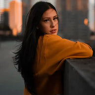 Frau mit langen dunklen Haaren, gelbem Pullover, Hoodie, auf Dach mit Stadt im Hintergrund