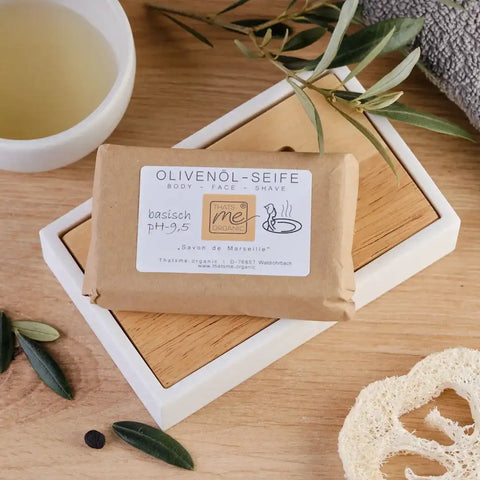 Natürliche Olivenölseife auf Holzseifenablage, umgeben von Olivenzweigen und Loofah.