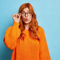 Frau mit roten Haaren und Brille im orangefarbenen Strickpullover vor blauem Hintergrund