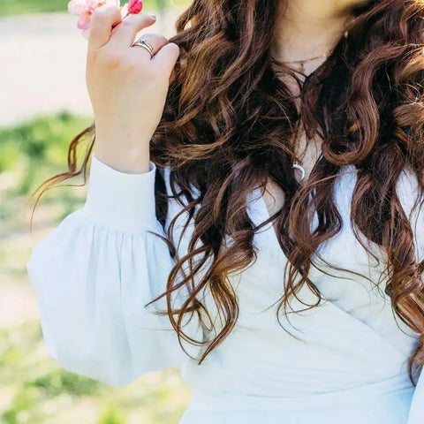 Frau mit langen braunen Locken und weißem Kleid hält rosa Blüten im Grünen