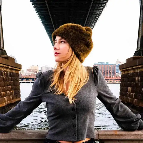 Frau mit brauner Fellmütze und grauer Bluse unter Brücke in urbaner Stadtlandschaft